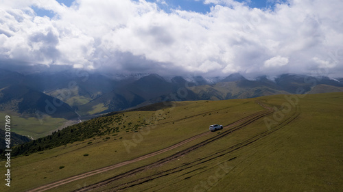 A white SUV is driving in the mountains through the fields. Aerial view from a drone of huge white clouds, snowy mountain peaks and green fields. Dirty off-road with puddles. Splashes from the wheels