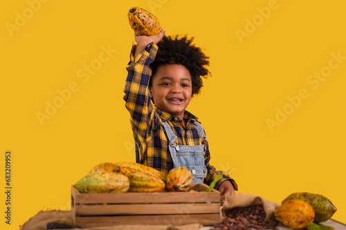 Portrait of happy African child boy farmer with afro hairstyle standing smiling and holding fresh cacao fruit on hand isolated on yellow background. photo