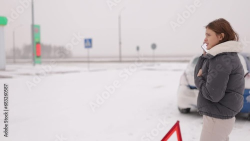 young female driver who crashed her car on a slippery winter road in the snow calls a tow truck with an emergency triangle next to her near the car.