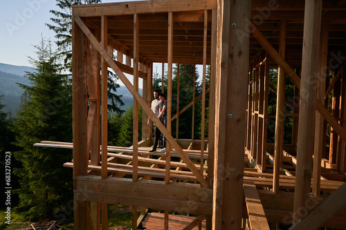 Man and woman examining their future wooden frame dwelling nestled in the mountains near forest. Youthful couple at construction site in early morning. Concept of contemporary ecological construction.