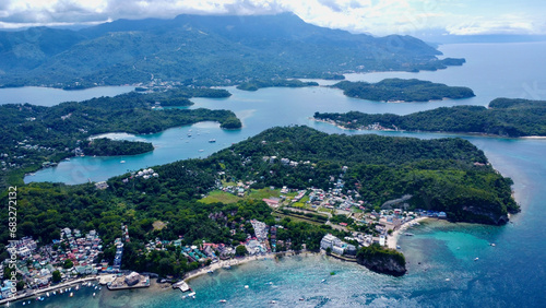 Top view of a small town on the shore of a lagoon on a tropical island. Aerial view of a coastal town among green forest on the shore of the blue sea. Sabang, Puerto Galera, Philippines. © Houston