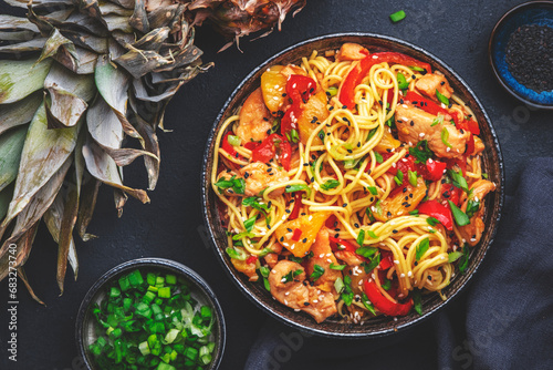 Stir fry noodles with chicken slices, pineapple, red paprika, chives, soy sauce and sesame seeds in ceramic bowl. Asian cuisine dish. Black table background, top view