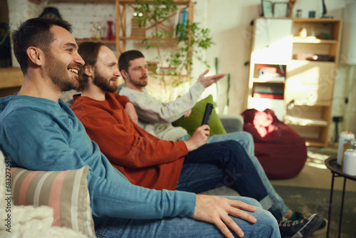 group of friends, men talking and having fun while watching TV sitting on sofa in living room at home. Concept of friendship.
