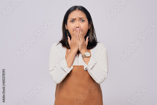 Surprised young Asian woman barista barman employee wearing brown apron working in coffee shop, posing covered mouth with hand isolated on white background. Small business startup concept