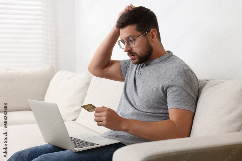 Upset man with credit card and laptop on sofa at home. Be careful - fraud Upset man with credit card and laptop on sofa at home. Be careful - fraud