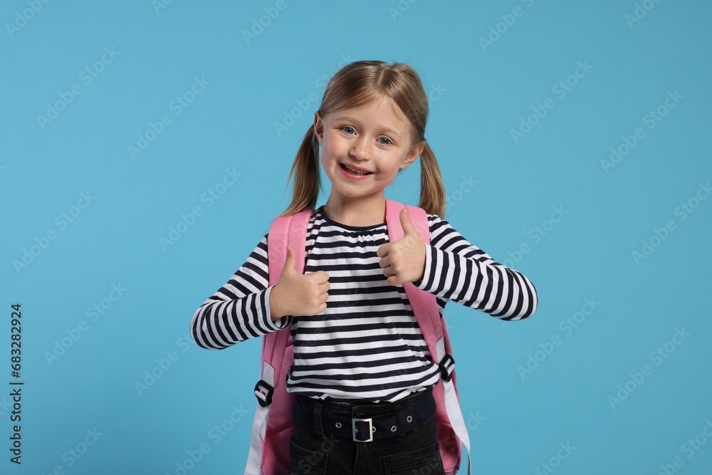 Happy schoolgirl with backpack showing thumbs up gesture on light blue background Happy schoolgirl with backpack showing thumbs up gesture on light blue background