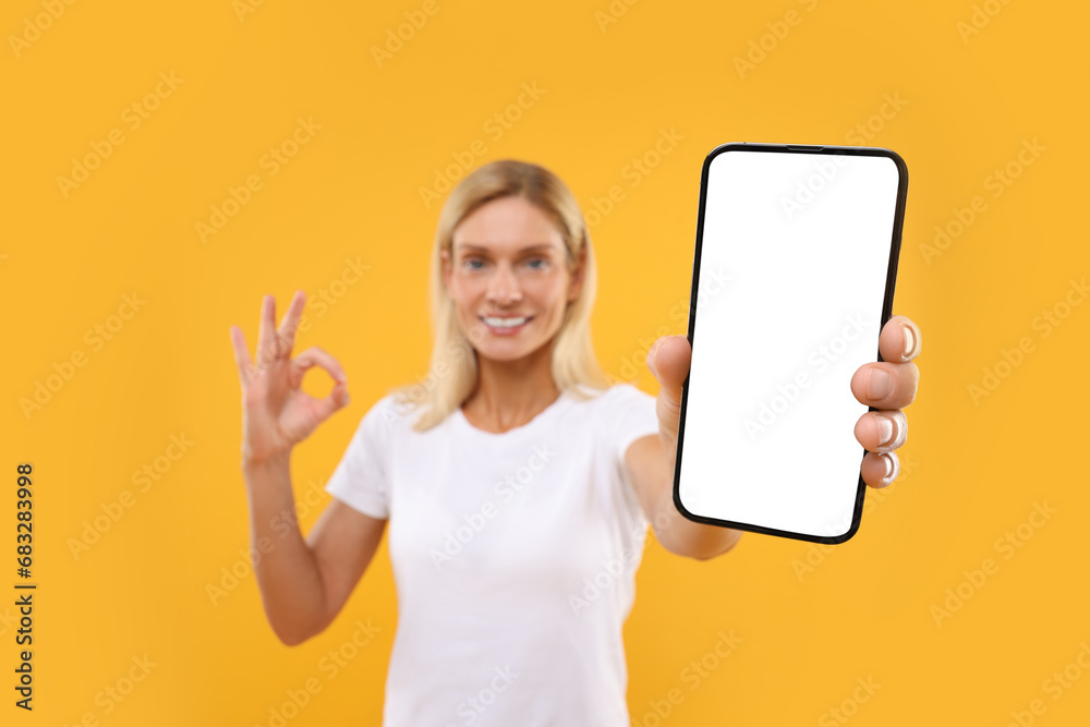 Happy woman holding smartphone with blank screen and showing OK gesture on orange background, selective focus