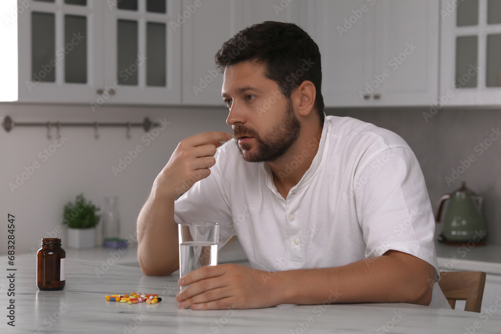 Depressed man with glass of water taking antidepressant pill at table in kitchen Depressed man with glass of water taking antidepressant pill at table in kitchen