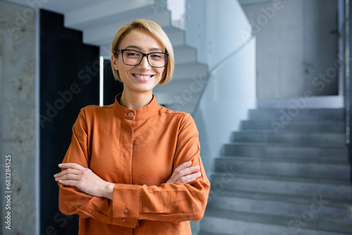 Portrait of young beautiful woman at workplace inside office, blonde business woman with crossed arms and glasses smiling and looking at camera, female worker standing near window.