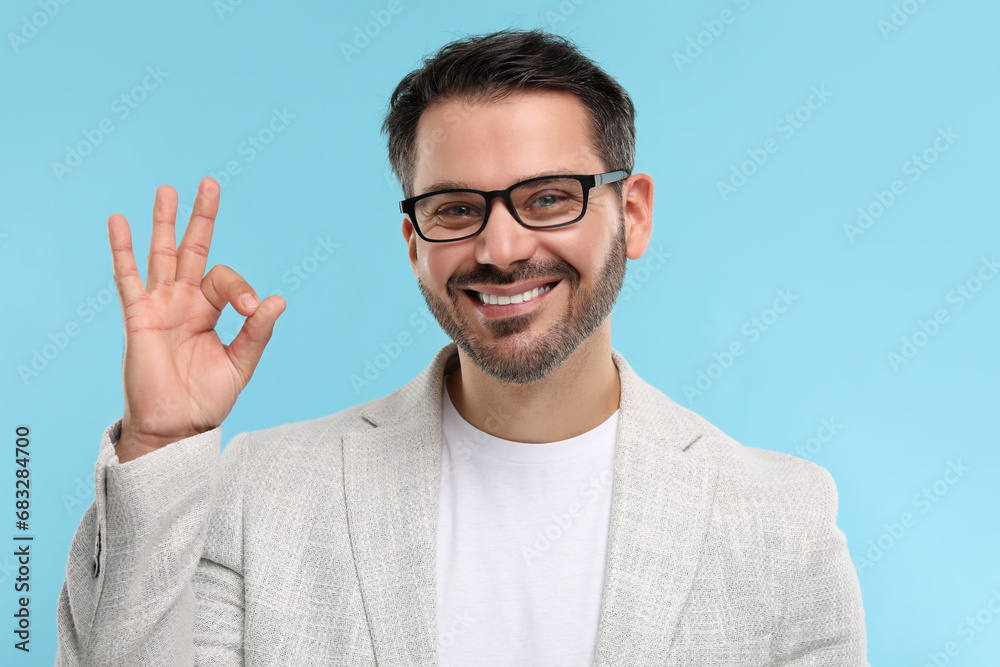 Portrait of happy man in stylish glasses showing OK gesture on light blue background Portrait of happy man in stylish glasses showing OK gesture on light blue background