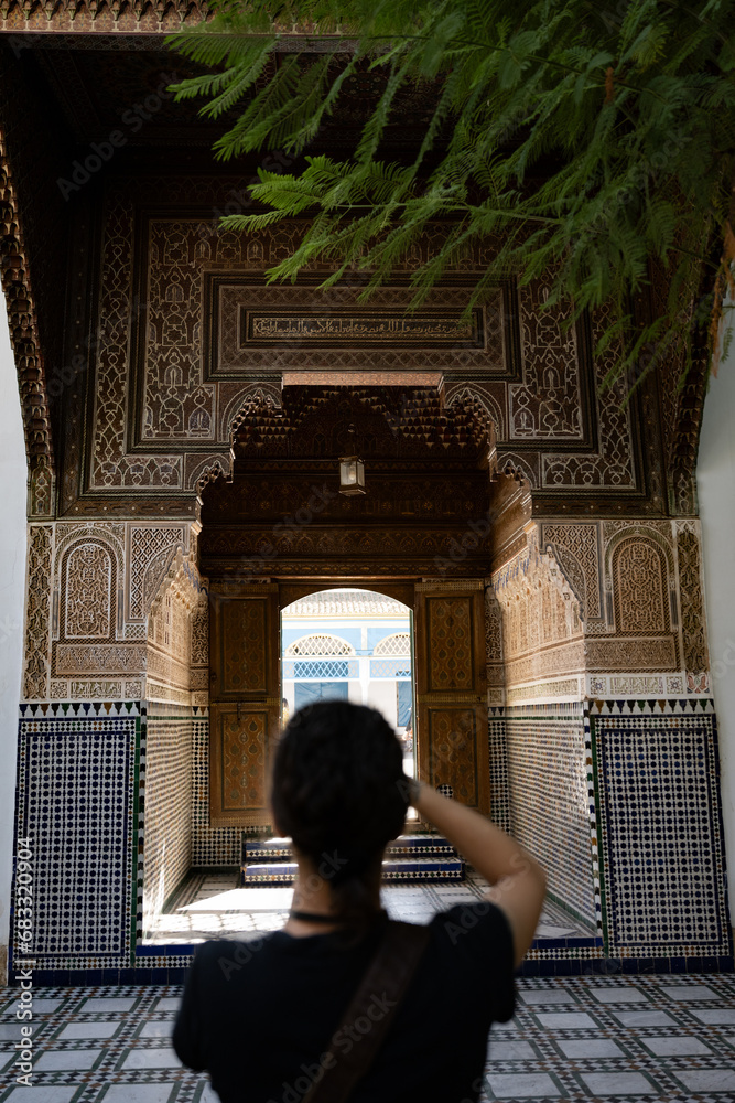 (Selective focus) Defocused girl in the foreground taking a photo at the Bahia Palace during a sunny day. The Bahia Palace in Marrakech is an impressive example of Moroccan architecture and design.