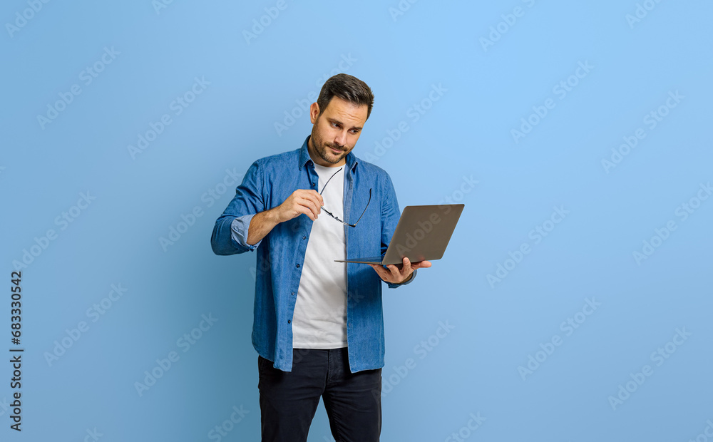 Confident young businessman holding eyeglasses and looking at laptop isolated on blue background Confident young businessman holding eyeglasses and looking at laptop isolated on blue background