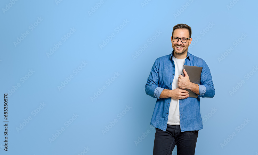 Confident young businessman holding digital tablet and smiling at camera against blue background - obrazy, fototapety, plakaty 