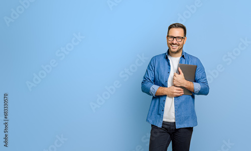 Confident young businessman holding digital tablet and smiling at camera against blue background