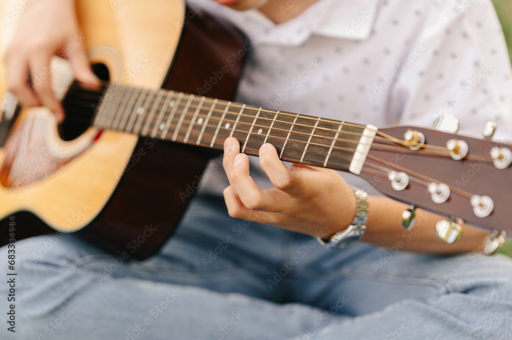 Close up shot of a teen boy learning to play acoustic guitar outdoors sitting in park.