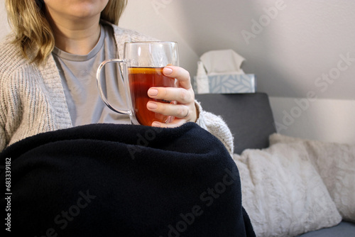 Sick woman sitting on sofa with blanket and hot tea with lemon and honey