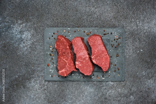 Three raw beef steaks on a black slate board on a dark concrete background. Steak ingredients, raw food, cooking.
