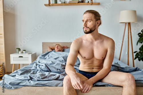upset bearded man in underpants sitting and looking away near love partner sleeping in bedroom