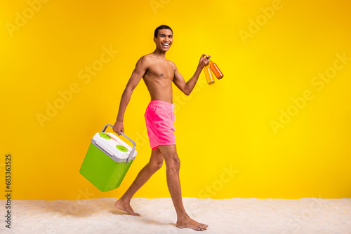 Full length photo of excited positive shirtless man holding refrigerator inviting drink beer empty space isolated yellow color background