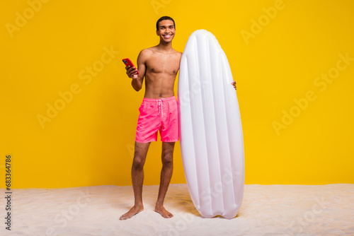 Full length photo of positive cheerful shirtless man holding floating matrass texting modern device isolated yellow color background