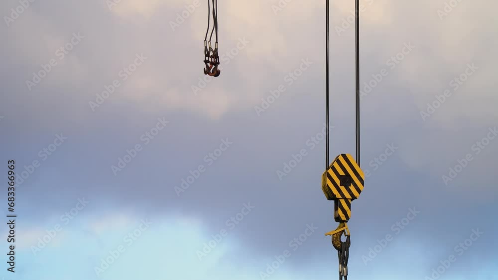 Steel cables and crane hooks in close-up against cloudy sky with flying bird on construction site. Construction site, yellow crane, warehouse shipyard Steel cables and crane hooks in close-up against cloudy sky with flying bird on construction site. Construction site, yellow crane, warehouse shipyard