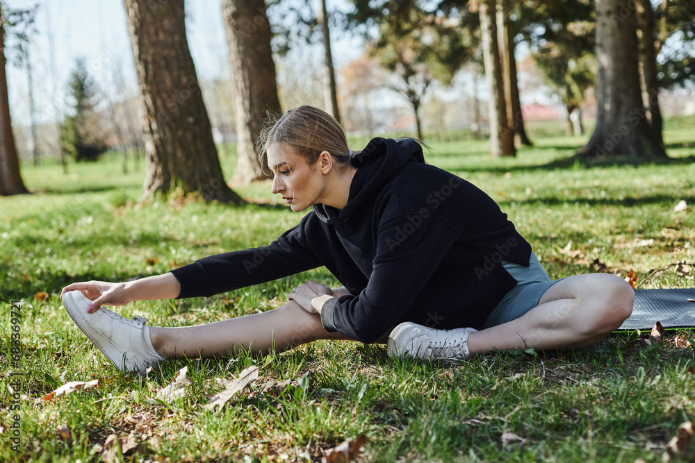 fit young woman with blonde hair and sportswear stretching while sitting on fitness mat in park fit young woman with blonde hair and sportswear stretching while sitting on fitness mat in park