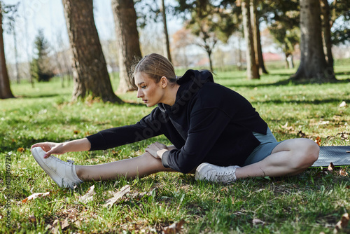 fit young woman with blonde hair and sportswear stretching while sitting on fitness mat in park