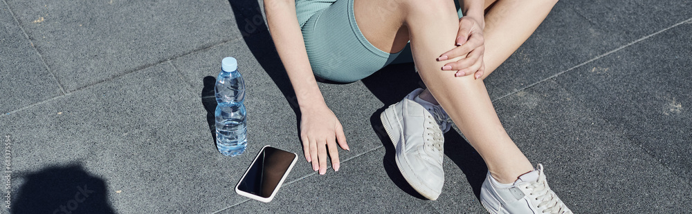 cropped banner of sportswoman in activewear sitting next to bottle of water and smartphone on floor