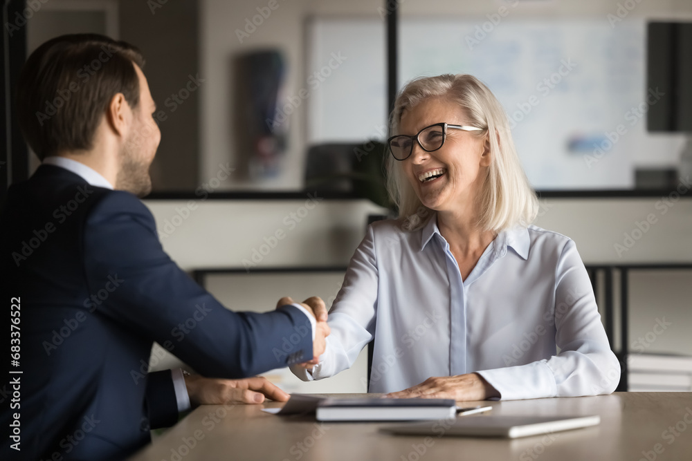 Cheerful elegant mature businesswoman shaking hands with younger business colleague man, laughing, ending meeting, starting collaboration, teamwork, giving handshake Cheerful elegant mature businesswoman shaking hands with younger business colleague man, laughing, ending meeting, starting collaboration, teamwork, giving handshake
