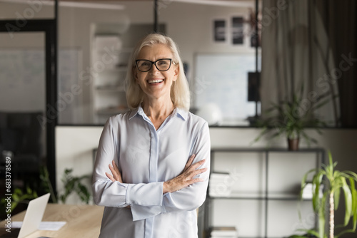 Cheerful confident old mature business woman in glasses posing in office with arms crossed, looking at camera, smiling, laughing, standing at workplace table. Professional head shot portrait photo