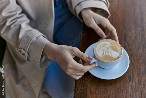 cropped view of young woman in trench coat holding cup with cappuccino in outdoor cafe, latte art