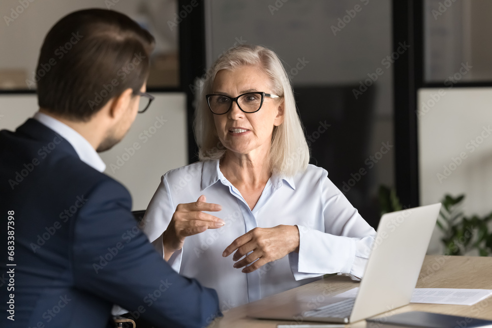 Serious elder professional woman talking to younger colleague man at workplace, working on project together, discussing collaboration, management strategy. Business partners speaking at office table - obrazy, fototapety, plakaty 