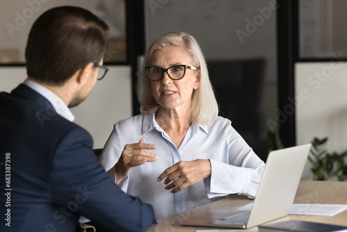 Serious elder professional woman talking to younger colleague man at workplace, working on project together, discussing collaboration, management strategy. Business partners speaking at office table