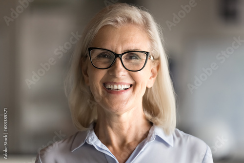 Positive mature elder business woman in stylish eyewear standing indoors, looking, smiling at camera with toothy smile, laughing. Female facial close up business portrait
