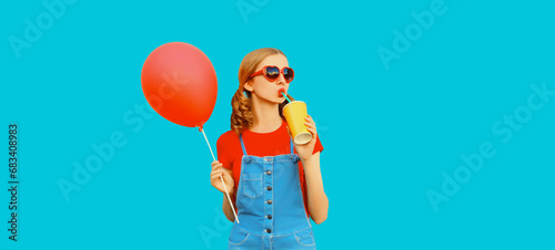 Happy cheerful young woman drinks fresh juice with balloon looking away wearing red heart shaped sunglasses, jumpsuit on blue background