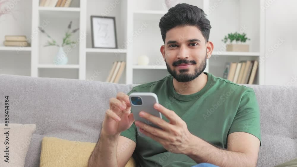 Portrait of handsome hindu guy in casual wear smiling and looking at camera while using modern smartphone on cozy sofa. Concept of technology, people and lifestyles. Portrait of handsome hindu guy in casual wear smiling and looking at camera while using modern smartphone on cozy sofa. Concept of technology, people and lifestyles.