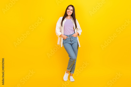 Full size photo of lovely girl wear white shirt denim trousers arms in pockets look empty promo space isolated on yellow color background