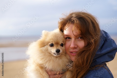 Happy young woman or teenager girl walking with her pet Pomeranian Spitz dog on the beach, hold puppy on hands. People love animals concept.