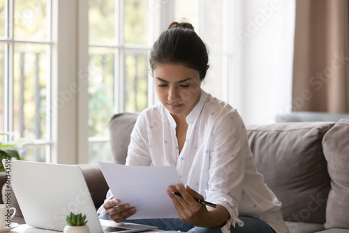 Young serious Indian woman housewife, entrepreneur sit on sofa holds paper document, learn agreement terms and conditions do paperwork at home looks attentive read formal important notice from bank photo