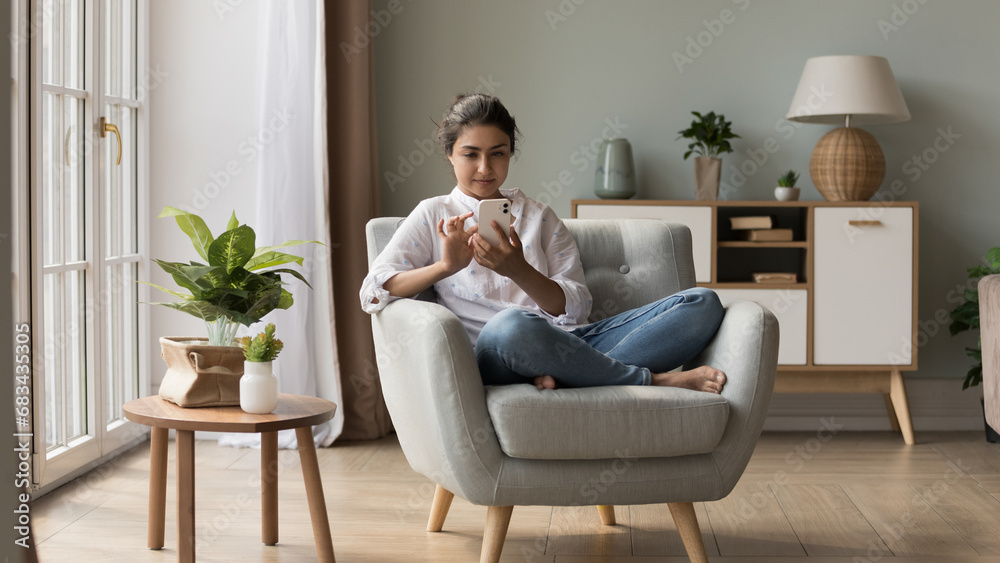 Attractive Indian woman relax on armchair in light fashionable living room holds smart phone chatting on-line, make order via internet electronic services. Leisure with tech, application usage concept Attractive Indian woman relax on armchair in light fashionable living room holds smart phone chatting on-line, make order via internet electronic services. Leisure with tech, application usage concept