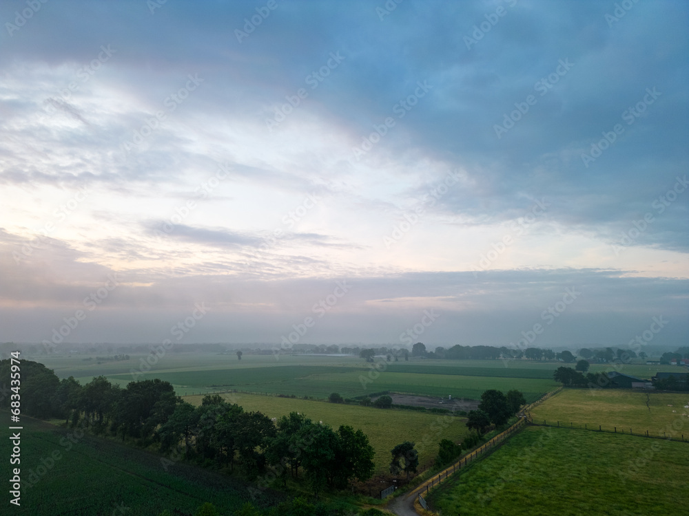 Fototapeta premium This image presents a tranquil early morning scene over the countryside, with the first light of dawn breaking across the horizon and casting a gentle glow over the landscape. The patchwork of fields