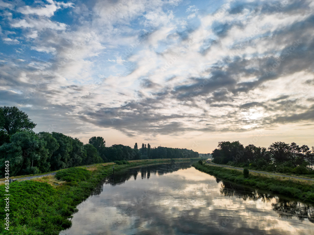 This photograph captures the peaceful essence of a river landscape at dawn. The still waters create a perfect mirror, reflecting the awakening sky adorned with streaks of clouds. The lush riverbanks