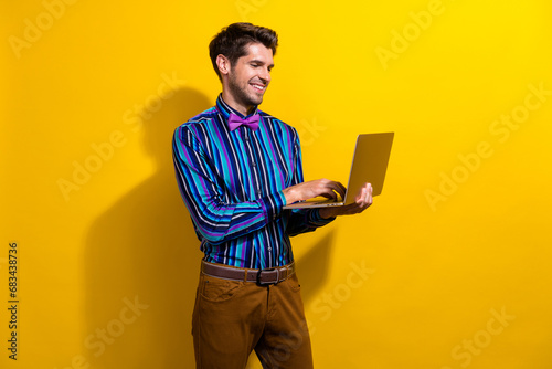 Portrait of focused positive guy dressed stylish shirt bow tie look at laptop chatting isolated on vibrant yellow color background