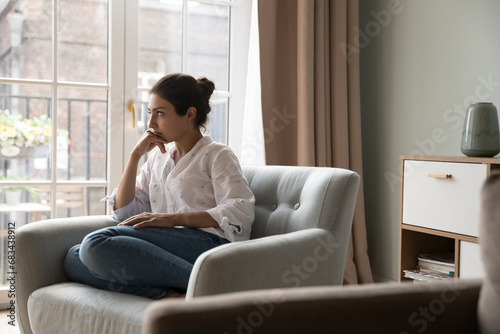 Worried young 25s Indian woman sit on armchair looks upset due to life concerns or break up, wait for boyfriend at home feels jealous, thinks over problems, search solution. Personal troubles concept