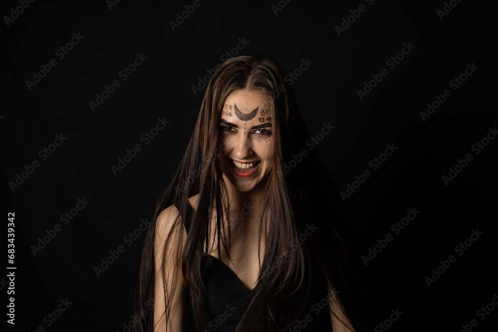 Evil laughing at the top of her voice, the witch looks into the camera on a black background in the twilight. On Halloween, a female witch grinning in a terrible grin looks at the camera.