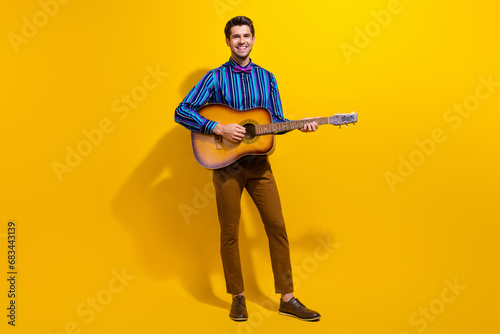 Full body photo of handsome cool man with bristle wear vintage bow tie standing playing song on guitar isolated on yellow color background © deagreez