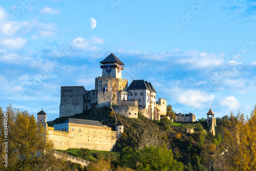 Trenčín Castle illuminated by the setting sun. In the background is a blue sky with clouds and a half moon. photo