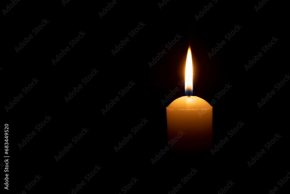 A single burning candle flame or light glowing on a big yellow candle on black or dark background on table in church for Christmas, funeral or memorial service with copy space A single burning candle flame or light glowing on a big yellow candle on black or dark background on table in church for Christmas, funeral or memorial service with copy space