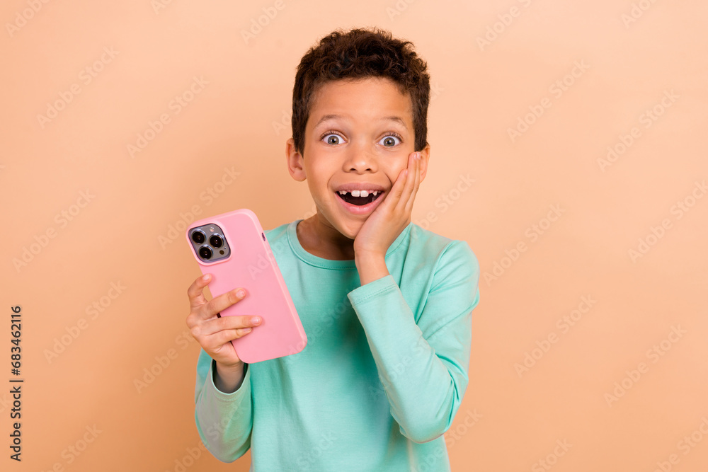 Photo of charming impressed cheerful boy wear trendy clothes playing videogame smartphone application isolated on beige color background Photo of charming impressed cheerful boy wear trendy clothes playing videogame smartphone application isolated on beige color background