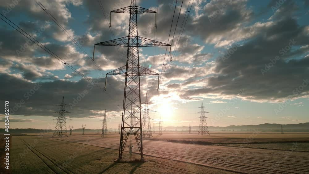 Electricity transmission towers with sunlit sky at sunrise aerial view. High voltage power pylon in field with nature landscape and cloudscape in morning drone shot